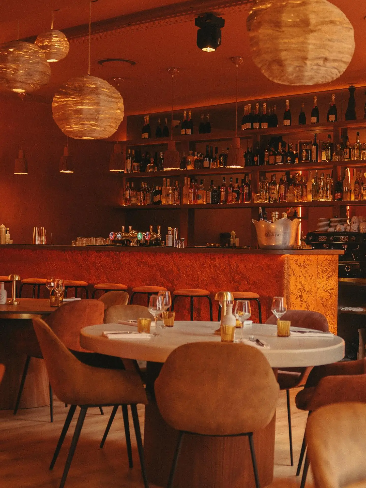 La salle du restaurant, tables rondes dressées et fauteuils en velours devant le bar et ses étagères de bouteilles