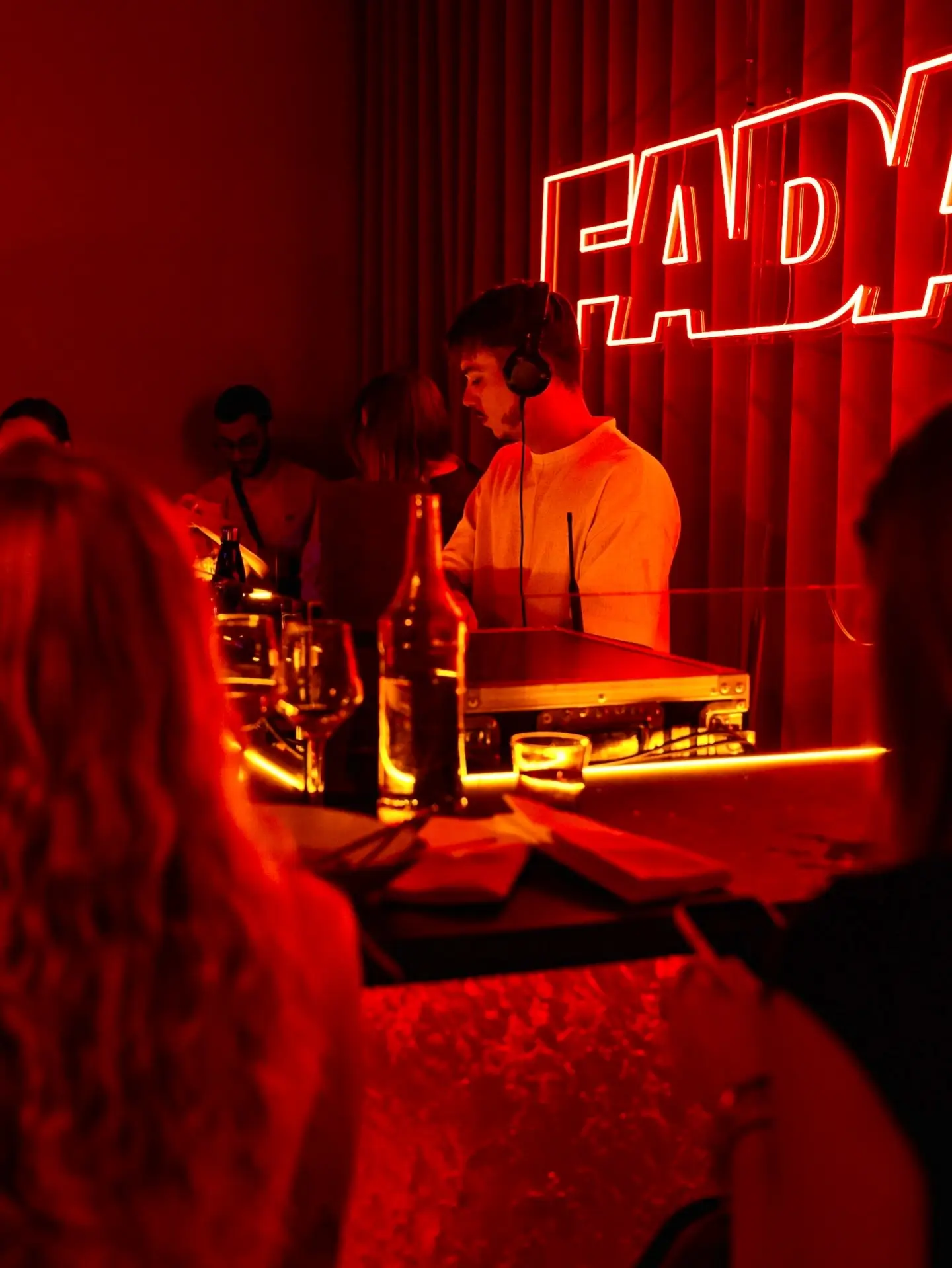 Un DJ aux platines sous l'enseigne néon FADA, dans la lumière rouge, des invités au premier plan